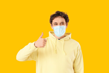 Young man in medical mask showing thumb-up on yellow background. Immunity concept