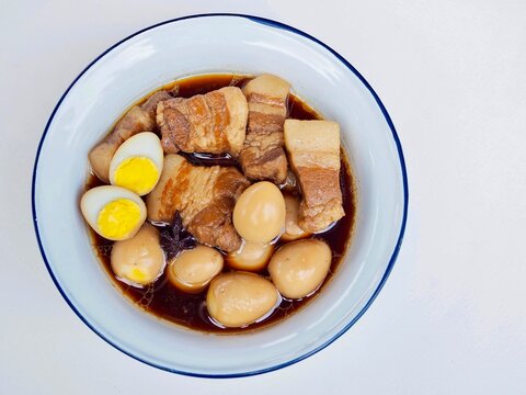 Boiled eggs in sweet broen soup with quail eggs and pork belly is served in a bowl on a white background. It's a Thai dish I made myself and it's very delicious.