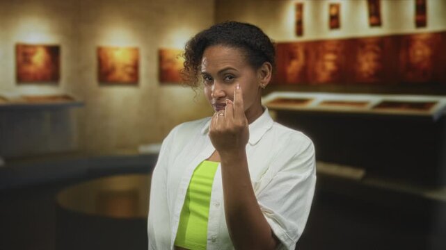 Woman in white shirt and neon green top points finger to camera inside a building gallery lined with framed art and display cases; confidence challenge assertion.