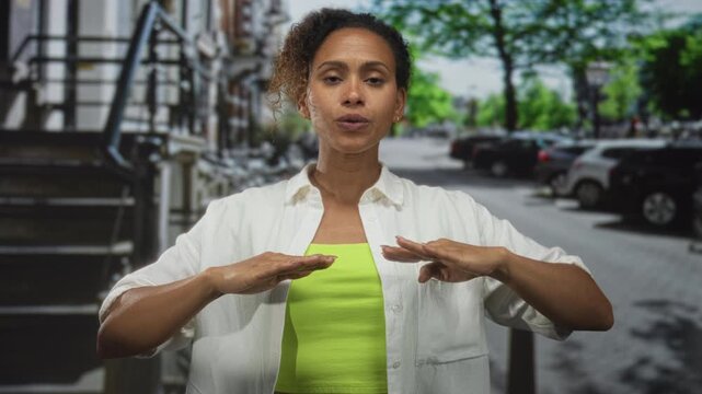 Woman with closed eyes and raised hands wearing neon green top and white shirt on a street lined with parked cars; calm concentration balance.