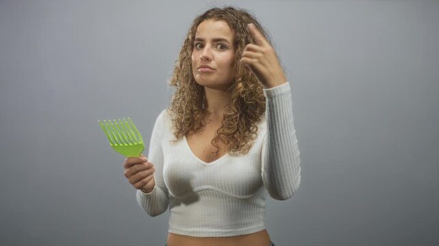 Woman with curly hair points finger while holding a green wide tooth comb and wearing a white crop top in a studio setting; confidence self care.