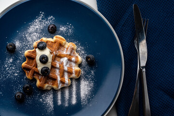 Belgian waffles or dessert with white cream and blueberries sprinkled with powdered sugar on a blue...