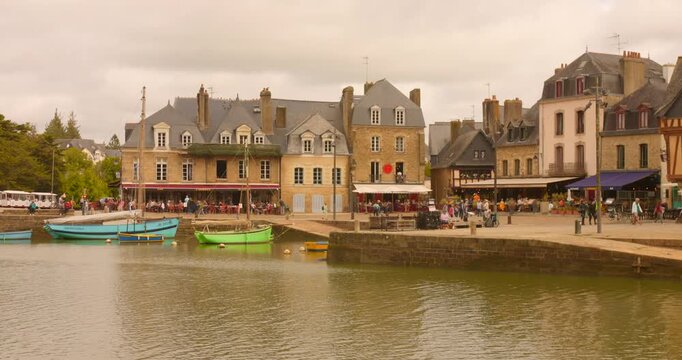 Footage shows Saint-Goustan Port in Auray, France with colorful boats in the water and historic buildings lining the shore. Midday lighting highlights the vibrant atmosphere and activity of the area.
