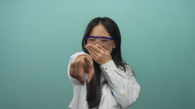 Scientist woman in a lab coat with glasses points playfully against an isolated blue background wall.
