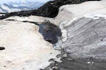 Fototapeta premium Water channel running through melting snow and dark rocks on Mount Elbrus