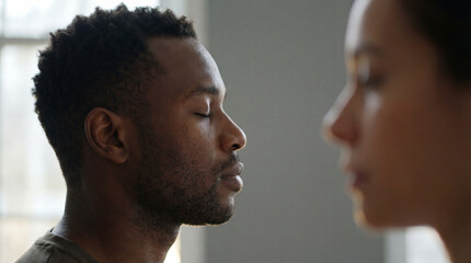 calm young man meditating with eyes closed indoors beside blurred figure, practicing mindfulness and relaxation in soft natural light