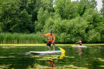 Adult paddler moving forward on a stand up paddle board in a calm lagoon at the Toronto Islands, Canada, during summer. Recreational skill progression with other participant in the background.