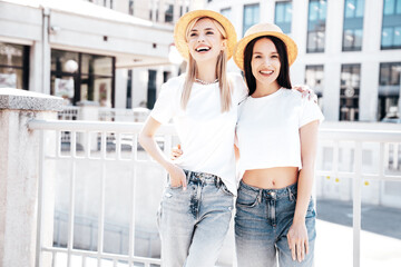Two young beautiful smiling hipster female in trendy summer white t-shirt and jeans clothes. Carefree women posing in street. Positive models having fun. Cheerful and happy. In hat