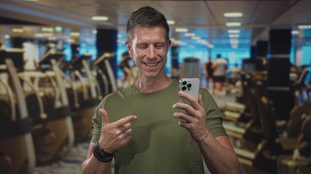 Man points finger to smartphone in building gym while smiling and showing hand; confidence connection.