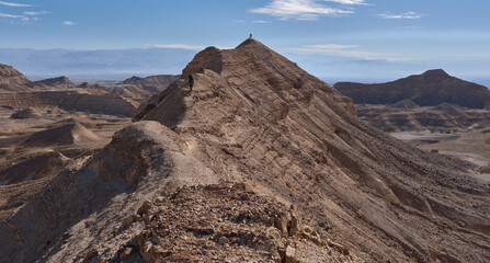 A sharp mountain ridge rises above wide and open valley of Nahal Zinin or Zanin in the Negev. A pair of tourists stands on a cliff with a panoramic view of the valley. Panoramic desert landscape. © A.Pushkin