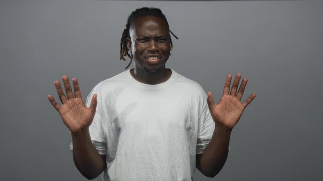 Young african american man in white t shirt with palms up and hands raised in a gray studio; disgust avoidance hesitation.