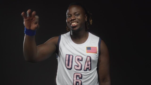 Young african american man in usa basketball jersey waving hand in black studio with direct gaze; quiet confidence.
