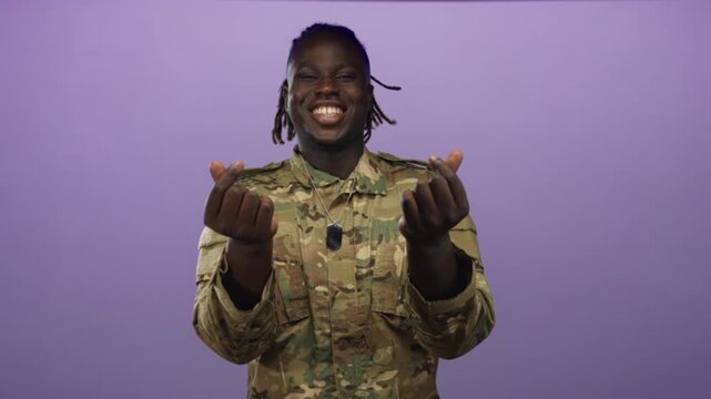 Man in army uniform smiling and making finger heart gesture with hands and dog tag in purple studio; pride duty.