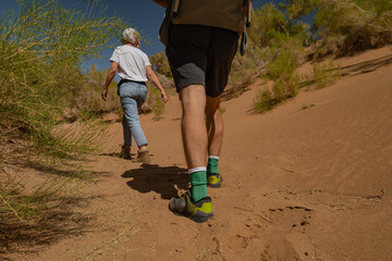 A couple hikes through a sandy canyon in Mongolia, where patches of rare green vegetation grow. Low rear-angle medium shot captures their boot soles and footprints.