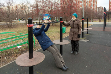 An elderly woman learns to exercise on outdoor exercise equipment with the help of another woman on a cool day. Active retirement.