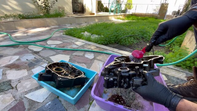 DIY mechanic cleaning very dirty engine crankcase parts on the ground outdoors, using small plastic basins, brush and gloves to remove heavy grease during home maintenance