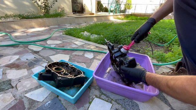 DIY mechanic cleaning very dirty engine crankcase parts on the ground outdoors, using small plastic basins, brush and gloves to remove heavy grease during home maintenance