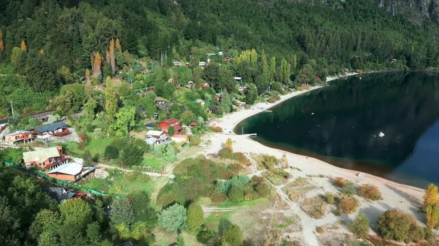 Forward drone aerial of lakefront village with colorful houses, sandy beach, autumn foliage, lush forest, and Ranco Lake shoreline