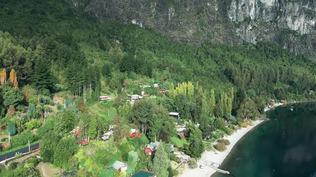 Revealing drone aerial of lakefront village with colorful houses, lush forest, dramatic rocky cliffs backdrop, and Ranco Lake shoreline