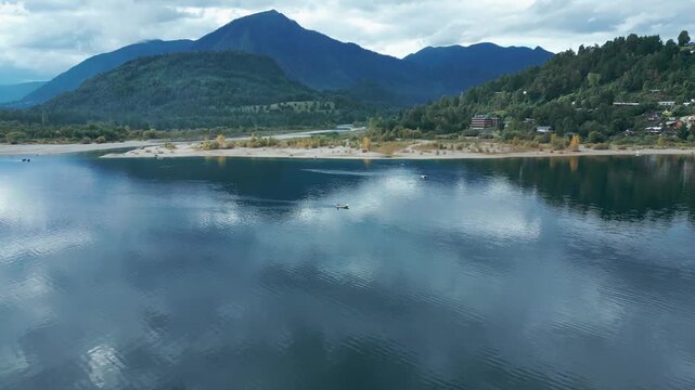 Tilting down drone aerial of boats on Ranco Lake with cloud reflections, mountains backdrop, beach, and serene Chilean landscape