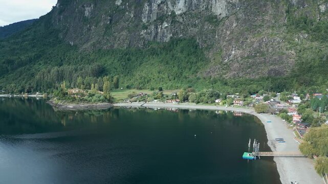 Backward drone aerial of Ranco Lake sandy beach with village houses, dramatic rocky cliff backdrop, and pristine Chilean shoreline