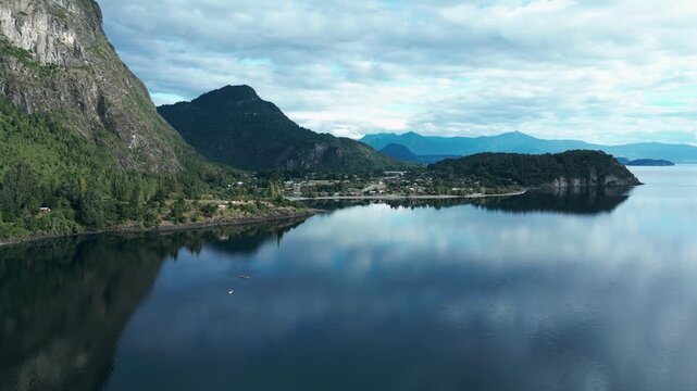 Panning drone aerial of Ranco Lake with mirror reflections, dramatic cliffs, mountains backdrop, peninsula, and Chilean village