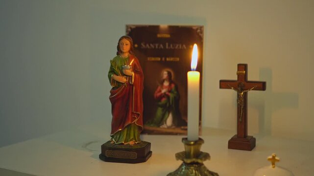 Religious setup with a statue of Saint Lucy a devotional booklet wooden crucifix and a burning candle on a table with warm light creating a sacred atmosphere for Christian prayer