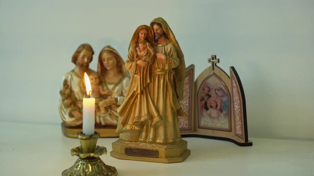 A collection of Holy Family religious statues and a small icon triptych arranged with a lit candle creating a prayerful and devotional atmosphere for Christian worship