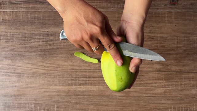 Top view video of hands peeling green mango with knife on wooden table
