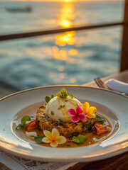 Gourmet dessert with grilled pineapple and cream scoop, garnished with frangipani flowers on a white plate. Tropical seaside dining at sunset with a beautiful golden hour ocean background.
