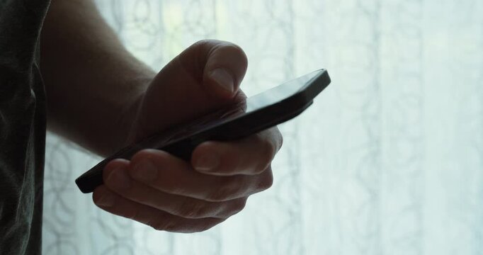 Close-up of a hand holding a smartphone and swiping on the screen in front of a bright window&mdash;ideal for TikTok scrolling, mobile addiction, doomscrolling, and digital lifestyle themes.