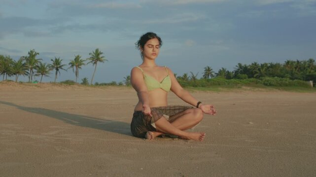 Woman sits in lotus postion for yoga meditation on a sandy beach at sunset, barefoot and serene, practicing mindfulness and balance while connecting with nature and the ocean horizon