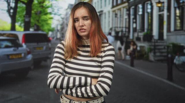 Woman with crossed arms in striped crop top showing midriff on street with parked cars and row buildings; annoyance defiance.