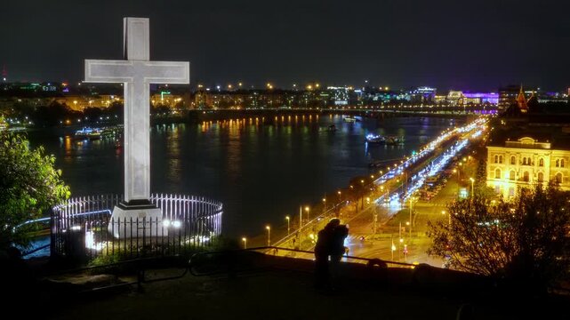 Romantic couple embracing at night on Gellert Hill in Budapest, overlooking the illuminated city skyline, Danube river, and a glowing stone cross under a dark sky. 