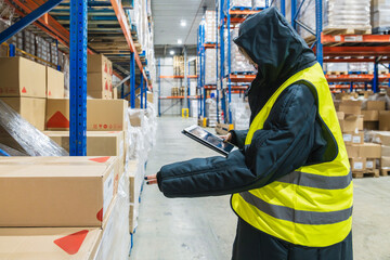 Worker in protective clothing using a tablet to inspect cardboard boxes stocked on shelves in a low temperature logistics warehouse