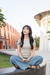 Pretty asian woman holding phone and looking away while sitting folding legs on bench in park plaza.