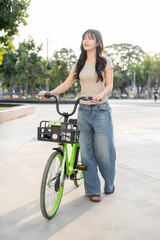Pretty asian woman walking with green rental bicycle on pavement in park or plaza under sunlight.