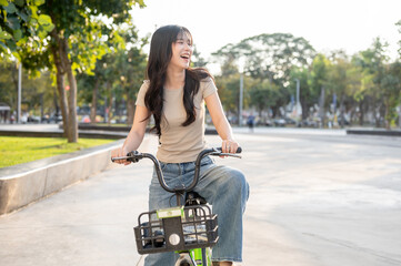 Laughing asian woman riding on a green rental bicycle on pavement in park or plaza under sunlight.