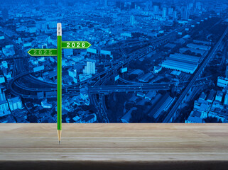 2026 and 2025 direction sign plate with green pencil on wooden table over modern office city tower, street, expressway and skyscraper, Business happy new year 2026 planning and strategy concept