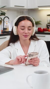 Young woman at home wears wireless headphones types message on smartphone takes break from tasks. Caucasian girl on sofa sits at table with laptop checks social app replies to chat relaxes happy mood