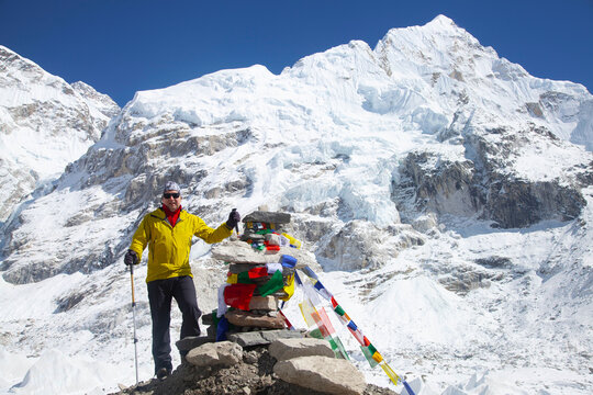 Adventurer in bright yellow jacket stands proudly beside colorful prayer flags at a stone monument, surrounded by majestic snow-capped peaks in a breathtaking mountain landscape.