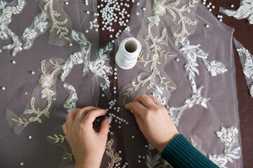close-up of female hands of seamstress tailor sewing wedding dress using beads and lace in the studio