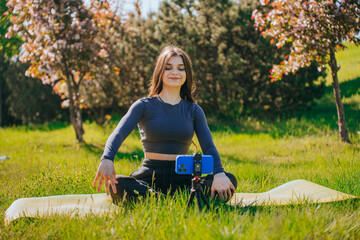 A woman sits cross-legged on a yoga mat, smiling contentedly, with lush greenery and blooming trees in the background.