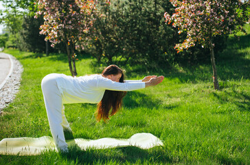 A woman in a white outfit performs a forward bend yoga pose on grass, framed by cherry blossoms and bathed in warm sunlight.