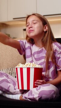 Teenager girl at night home watches film eats popcorn from bowl giggles then bursts into loud laugh. Caucasian child kid on sofa at evening points at screen claps hands enjoys fun story pure big joy