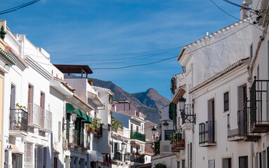 A narrow street lined with whitewashed houses and green awnings leads toward rugged mountains under a blue sky in Frigiliana, capturing the charm of a traditional Andalusian village in sunny Spain.