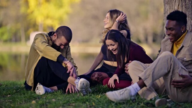 Happy young friends enjoy a lively conversation and shared laughter while relaxing on the grass by a tranquil lake. Their diverse group finds joy and connection in the beautiful autumn park setting.