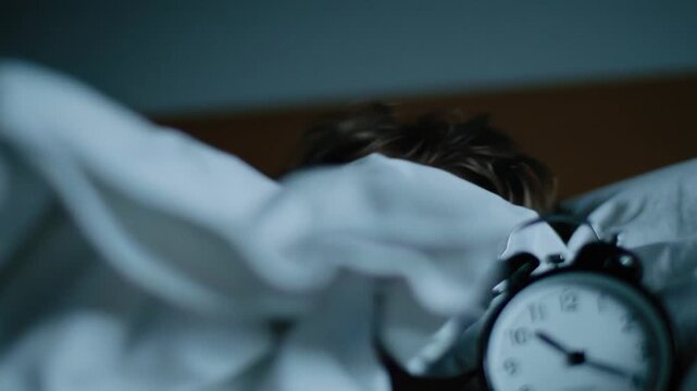 A young child slowly awakens in the early morning, peering curiously from a blanket. An alarm clock sits nearby, marking the start of a new day