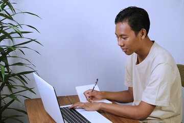 Young Asian Gen Z man studying online and taking notes while working on a laptop at home. Authentic daily life, education, productivity, and remote learning lifestyle concept.