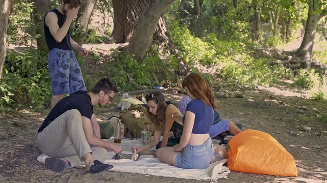 A diverse group of young friends gathers for a relaxing picnic in a lush green park, playing a board game and enjoying each other's company on a beautiful summer day.